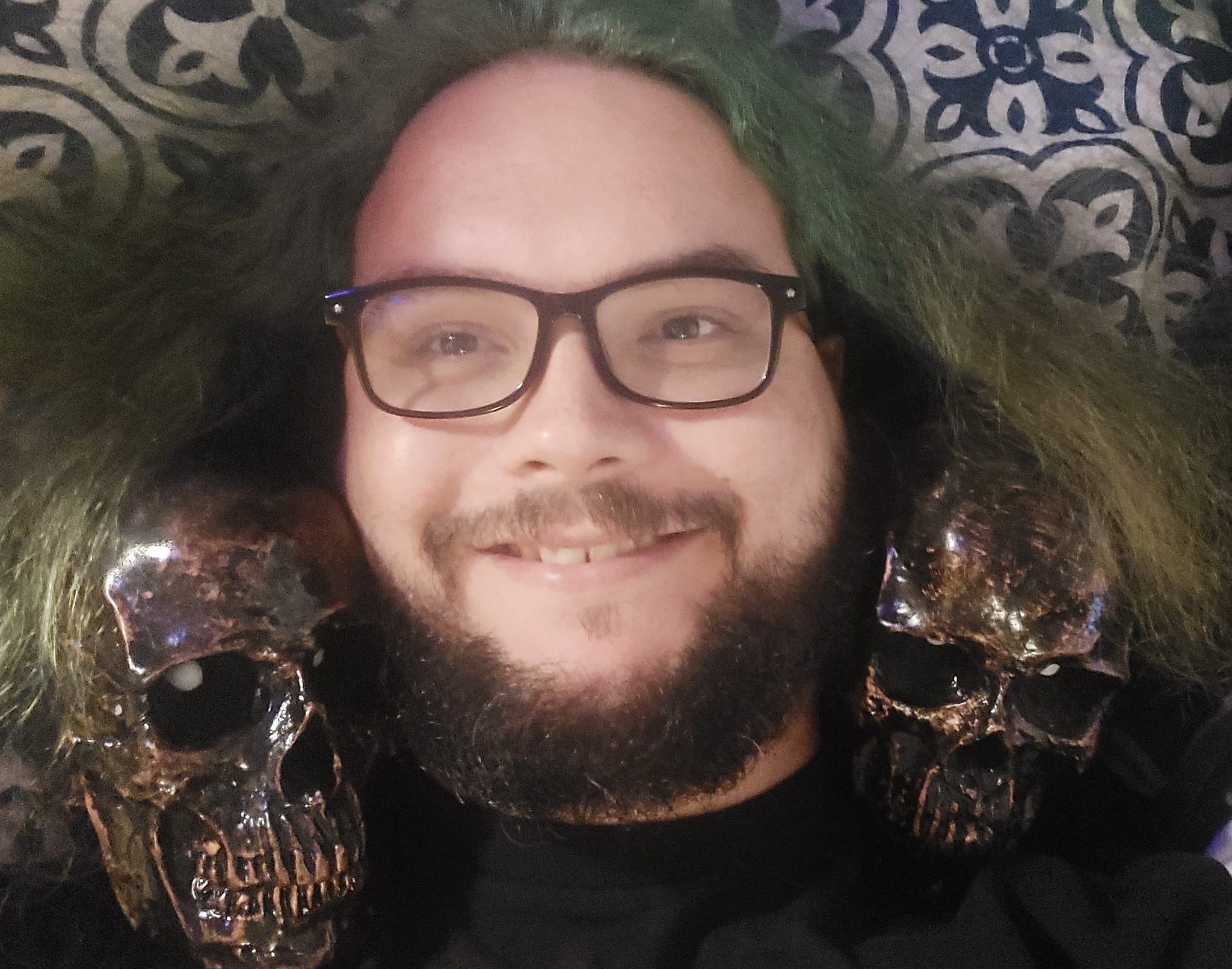 A man with green hair and a beard lays in bed with two plastic halloween skulls on his sholders